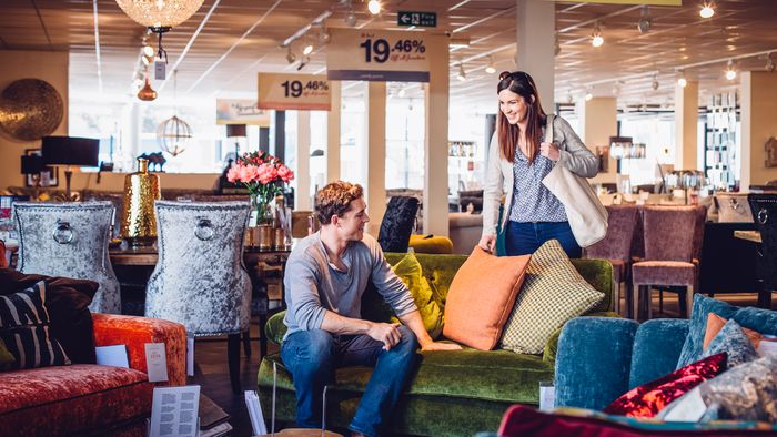 A young couple shops at a furniture store, testing out a green, velvet loveseat.